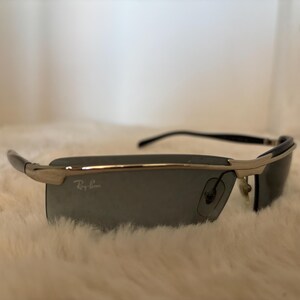 May include: Silver and black sunglasses with dark gray lenses. The sunglasses have a rectangular shape and the Ray-Ban logo is visible on the lens. The frame is a combination of silver and black.