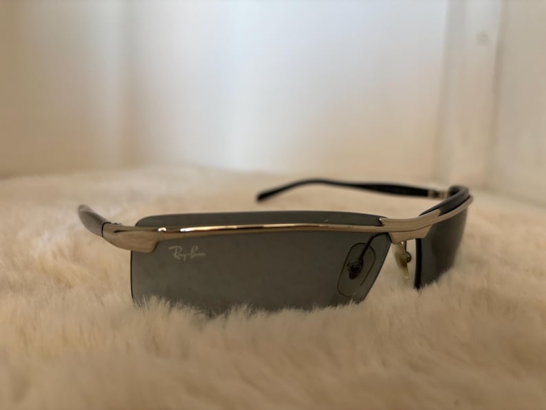 May include: Silver and black sunglasses with dark gray lenses. The sunglasses have a rectangular shape and the Ray-Ban logo is visible on the lens. The frame is a combination of silver and black.
