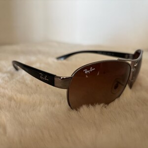 May include: Sunglasses with a brown gradient lens and black arms. The sunglasses have a silver frame and the Ray-Ban logo is visible on the lens and arm.
