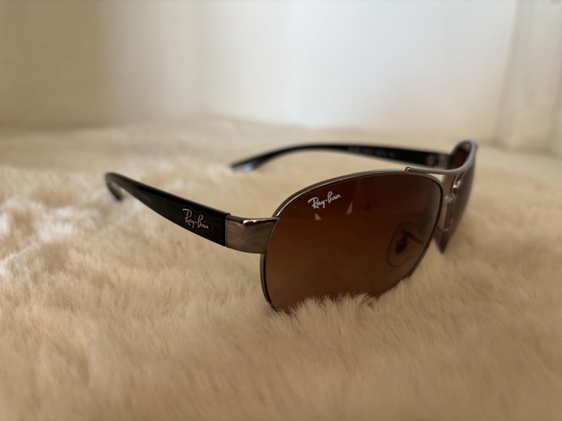 May include: Sunglasses with a brown gradient lens and black arms. The sunglasses have a silver frame and the Ray-Ban logo is visible on the lens and arm.