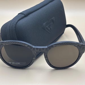 May include: Dark blue sunglasses with a geometric pattern on the frames, in front of a black case. The lenses are a gradient brown colour. The sunglasses have the word "MELLOW" printed on the lens.