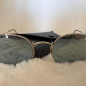 K&ouml;nnte beinhalten: Silberfarbene Sonnenbrille mit verspiegelten Gl&auml;sern. Die runden Gl&auml;ser reflektieren einen weichen, hellfarbigen Hintergrund. Die Sonnenbrille liegt auf einer wei&szlig;en, flauschigen Oberfl&auml;che, mit einem schwarzen Etui dahinter.