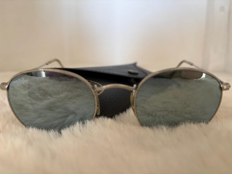K&ouml;nnte beinhalten: Silberfarbene Sonnenbrille mit verspiegelten Gl&auml;sern. Die runden Gl&auml;ser reflektieren einen weichen, hellfarbigen Hintergrund. Die Sonnenbrille liegt auf einer wei&szlig;en, flauschigen Oberfl&auml;che, mit einem schwarzen Etui dahinter.