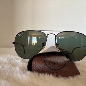 Pode incluir: &Oacute;culos de sol aviador pretos com lentes verdes repousam sobre um estojo de couro castanho. Os &oacute;culos de sol t&ecirc;m uma ponte dupla e o logotipo da Ray-Ban &eacute; vis&iacute;vel na lente superior esquerda. O estojo tem um logotipo em relevo.