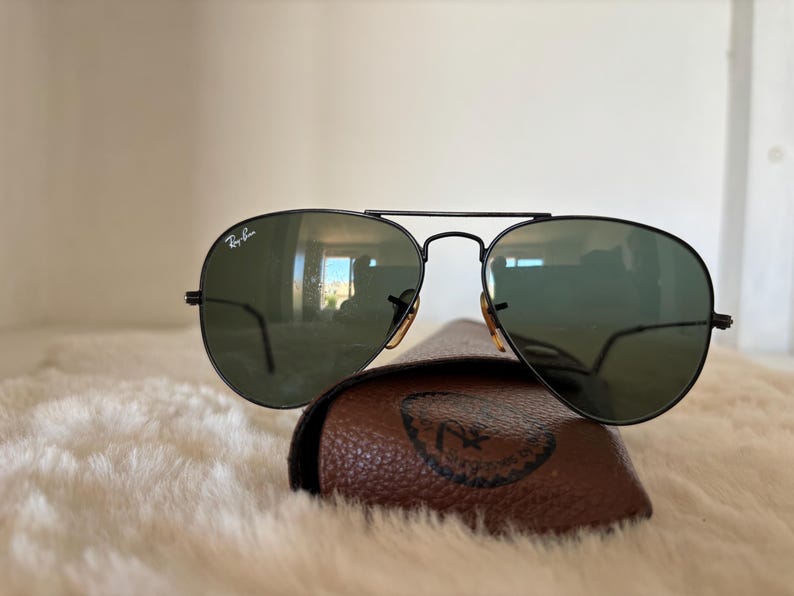 Pode incluir: &Oacute;culos de sol aviador pretos com lentes verdes repousam sobre um estojo de couro castanho. Os &oacute;culos de sol t&ecirc;m uma ponte dupla e o logotipo da Ray-Ban &eacute; vis&iacute;vel na lente superior esquerda. O estojo tem um logotipo em relevo.