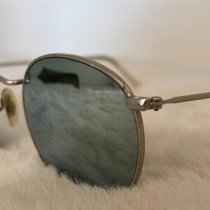 K&ouml;nnte beinhalten: Nahaufnahme einer silberfarbenen Sonnenbrille mit runden Gl&auml;sern. Die Gl&auml;ser haben eine reflektierende, blau-gr&uuml;ne T&ouml;nung. Die B&uuml;gel der Brille sind d&uuml;nn und silberfarben, und die Nasenpads sind hellbeige. Die Brille ist vor einem weichen, wei&szlig;en Hintergrund platziert.