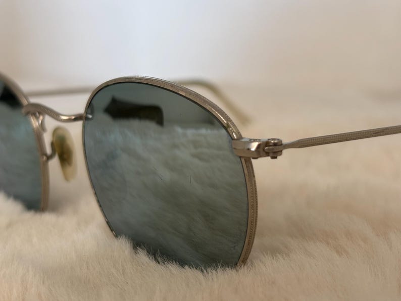 K&ouml;nnte beinhalten: Nahaufnahme einer silberfarbenen Sonnenbrille mit runden Gl&auml;sern. Die Gl&auml;ser haben eine reflektierende, blau-gr&uuml;ne T&ouml;nung. Die B&uuml;gel der Brille sind d&uuml;nn und silberfarben, und die Nasenpads sind hellbeige. Die Brille ist vor einem weichen, wei&szlig;en Hintergrund platziert.