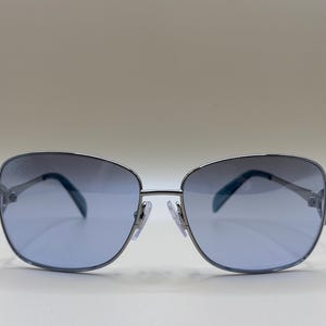 Peut inclure: Lunettes de soleil à monture argentée avec verres bleus dégradés. Les verres de forme ovale sont montés dans une monture en métal argenté avec un double pont et des plaquettes de nez. Les verres passent d'un bleu plus foncé en haut à un bleu plus clair en bas.