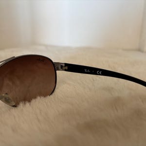 May include: Sunglasses with a dark metal frame and gradient brown lenses. The black temples have the brand name and "CE" printed on them. The sunglasses are resting on a white, fluffy surface.