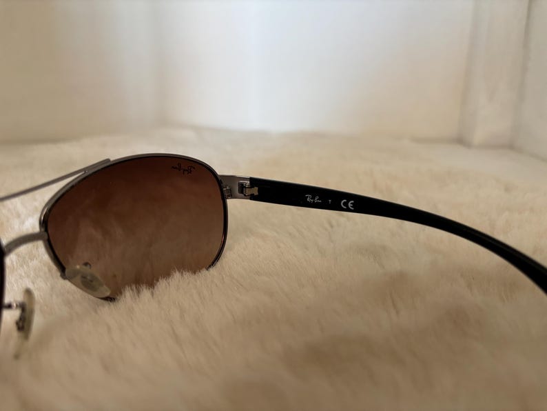 May include: Sunglasses with a dark metal frame and gradient brown lenses. The black temples have the brand name and "CE" printed on them. The sunglasses are resting on a white, fluffy surface.