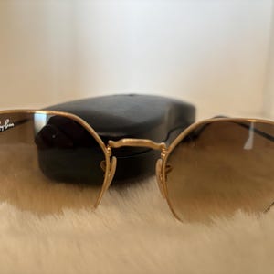 Puede incluir: Gafas de sol doradas con lentes degradadas marrones. El logotipo de Ray-Ban es visible en la lente izquierda. Un estuche negro descansa detrás de las gafas de sol sobre una superficie blanca y esponjosa.