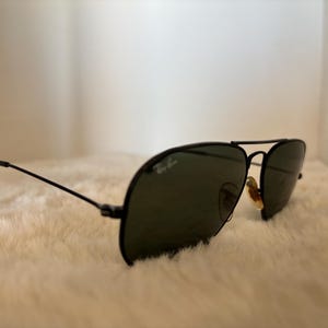 Pode incluir: &Oacute;culos de sol aviador pretos com lentes verde-escuras. Os &oacute;culos t&ecirc;m uma arma&ccedil;&atilde;o fina de metal preto e o logotipo da Ray-Ban &eacute; vis&iacute;vel na lente. Os &oacute;culos est&atilde;o sobre uma superf&iacute;cie branca e fofa.