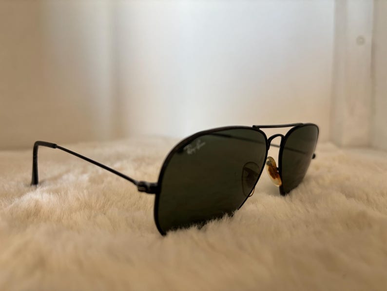 Pode incluir: &Oacute;culos de sol aviador pretos com lentes verde-escuras. Os &oacute;culos t&ecirc;m uma arma&ccedil;&atilde;o fina de metal preto e o logotipo da Ray-Ban &eacute; vis&iacute;vel na lente. Os &oacute;culos est&atilde;o sobre uma superf&iacute;cie branca e fofa.