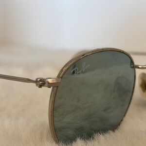 K&ouml;nnte beinhalten: Nahaufnahme einer Ray-Ban Sonnenbrille mit silbernen Rahmen und blau get&ouml;nten Gl&auml;sern. Das Ray-Ban-Logo ist auf dem Glas zu sehen. Die Brille hat eine klassische, runde Form und ist vor einem weichen, wei&szlig;en Hintergrund.