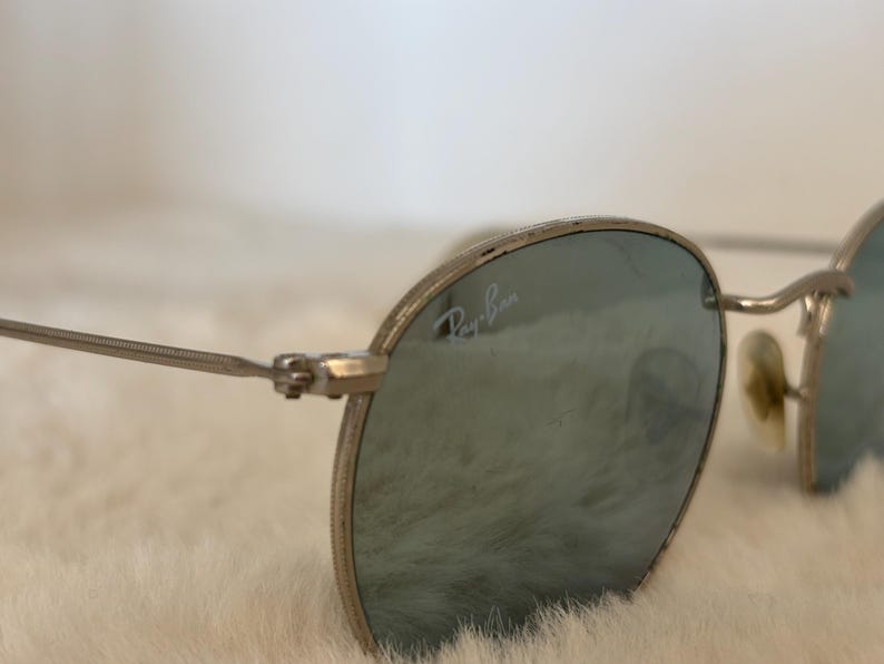 K&ouml;nnte beinhalten: Nahaufnahme einer Ray-Ban Sonnenbrille mit silbernen Rahmen und blau get&ouml;nten Gl&auml;sern. Das Ray-Ban-Logo ist auf dem Glas zu sehen. Die Brille hat eine klassische, runde Form und ist vor einem weichen, wei&szlig;en Hintergrund.