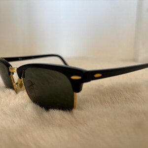 May include: Black sunglasses with a gold-coloured bridge and accents. The sunglasses have a classic design with a black frame and dark lenses. The glasses are resting on a white, fluffy surface.