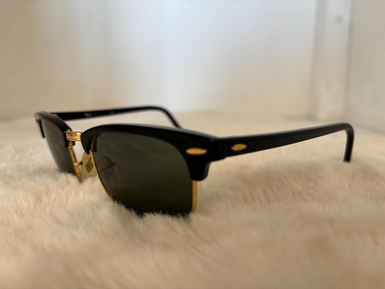 May include: Black sunglasses with a gold-coloured bridge and accents. The sunglasses have a classic design with a black frame and dark lenses. The glasses are resting on a white, fluffy surface.