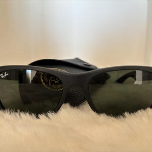 K&ouml;nnte beinhalten: Schwarze Sonnenbrille mit mattem Finish und klassischem rechteckigem Rahmen. Das Ray-Ban-Logo ist auf dem linken B&uuml;gel sichtbar. Die dunklen Gl&auml;ser liegen auf einer weichen, hellen Oberfl&auml;che, mit einem schwarzen Etui im Hintergrund.
