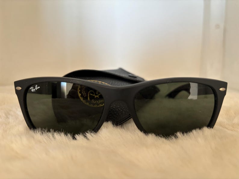 K&ouml;nnte beinhalten: Schwarze Sonnenbrille mit mattem Finish und klassischem rechteckigem Rahmen. Das Ray-Ban-Logo ist auf dem linken B&uuml;gel sichtbar. Die dunklen Gl&auml;ser liegen auf einer weichen, hellen Oberfl&auml;che, mit einem schwarzen Etui im Hintergrund.