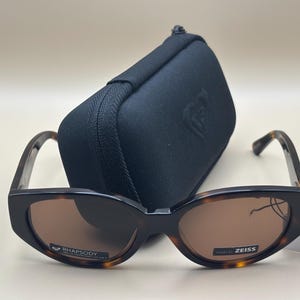 Può includere: Occhiali da sole con montatura nera e tartarugata e lenti marroni davanti a un astuccio nero. Sugli occhiali da sole sono stampate le scritte "RHAPSODY" e "ZEISS". È attaccata un'etichetta bianca con il logo del marchio.