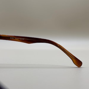 May include: Close-up of a brown and amber-colored eyeglass arm. The arm is curved and tapers to a rounded tip. The text "B&L RAY-BAN" is printed on the arm. The glasses are likely for vision correction or sun protection.