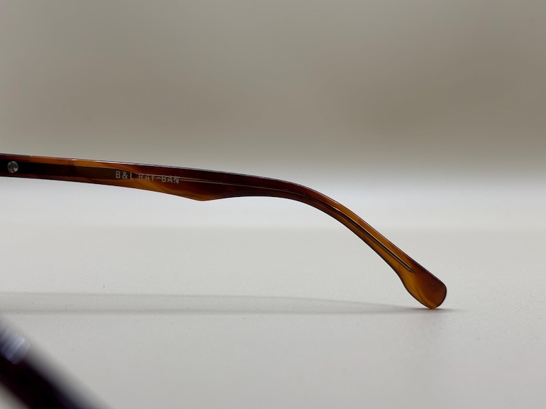 May include: Close-up of a brown and amber-colored eyeglass arm. The arm is curved and tapers to a rounded tip. The text "B&L RAY-BAN" is printed on the arm. The glasses are likely for vision correction or sun protection.