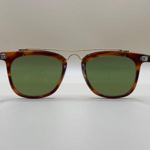 May include: Brown tortoiseshell sunglasses with green lenses and a gold-colored metal bridge. The sunglasses have a classic, retro design. The arms of the sunglasses are also brown tortoiseshell.