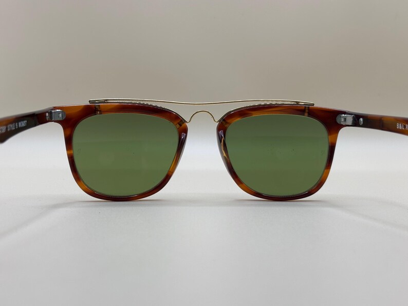 May include: Brown tortoiseshell sunglasses with green lenses and a gold-colored metal bridge. The sunglasses have a classic, retro design. The arms of the sunglasses are also brown tortoiseshell.