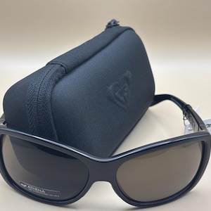 Può includere: Occhiali da sole neri con lenti marroni e custodia nera abbinata. Gli occhiali hanno una montatura spessa e un design curvo. La custodia è rettangolare con chiusura a zip e logo. Gli occhiali sono etichettati "ATHENA".