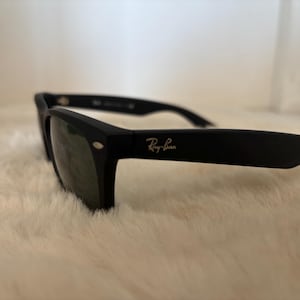 K&ouml;nnte beinhalten: Schwarze Sonnenbrille mit dem Ray-Ban-Logo in heller Farbe. Die Sonnenbrille hat einen klassischen rechteckigen Rahmen und dunkle Gl&auml;ser. Die Sonnenbrille liegt auf einer wei&szlig;en, flauschigen Oberfl&auml;che.