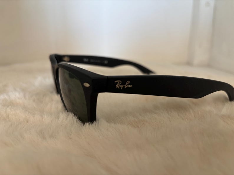 K&ouml;nnte beinhalten: Schwarze Sonnenbrille mit dem Ray-Ban-Logo in heller Farbe. Die Sonnenbrille hat einen klassischen rechteckigen Rahmen und dunkle Gl&auml;ser. Die Sonnenbrille liegt auf einer wei&szlig;en, flauschigen Oberfl&auml;che.