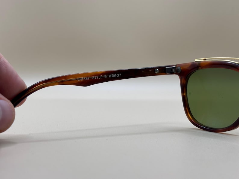 May include: Close-up of vintage sunglasses with a tortoiseshell frame and green lenses. The arm of the glasses has the text "GATSBY STYLE 5 W0937". The glasses have a gold-colored top bar.