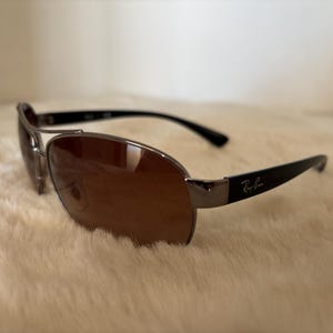 May include: Sunglasses with brown lenses and a silver frame. The temples are black and feature the Ray-Ban logo. The sunglasses are resting on a soft, cream-colored surface.
