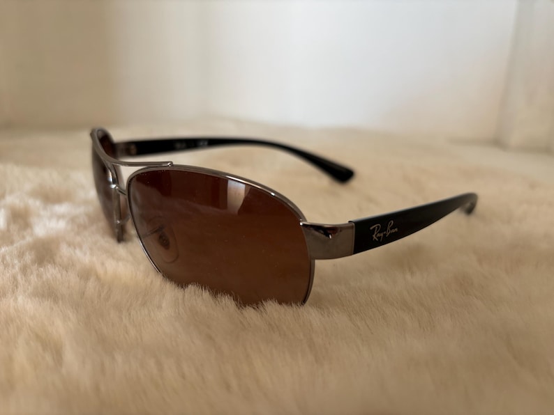 May include: Sunglasses with brown lenses and a silver frame. The temples are black and feature the Ray-Ban logo. The sunglasses are resting on a soft, cream-colored surface.