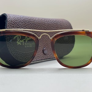 May include: Tortoiseshell sunglasses with green lenses and a gold-tone brow bar. The sunglasses are in a brown leather case. The Ray-Ban logo is visible on the lens.