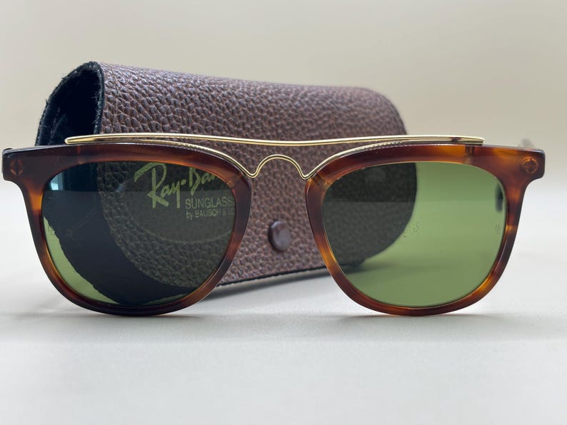 May include: Tortoiseshell sunglasses with green lenses and a gold-tone brow bar. The sunglasses are in a brown leather case. The Ray-Ban logo is visible on the lens.