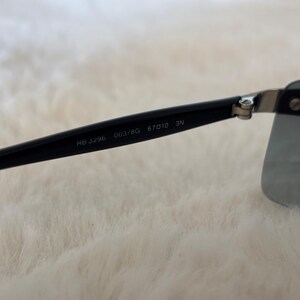 May include: Black sunglasses with a rectangular lens shape. The black arm of the sunglasses has the text "RB 3296 003/8G 67010 3N" printed on it. The sunglasses are set against a soft, white background.