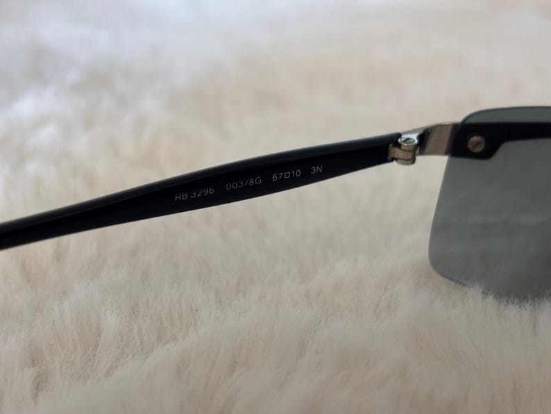 May include: Black sunglasses with a rectangular lens shape. The black arm of the sunglasses has the text "RB 3296 003/8G 67010 3N" printed on it. The sunglasses are set against a soft, white background.