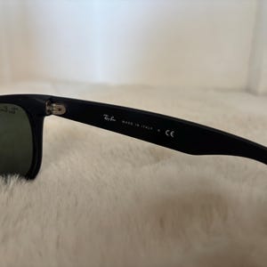 K&ouml;nnte beinhalten: Schwarze Sonnenbrille mit dunklen Gl&auml;sern. Der Rahmen ist schwarz mit dem Text "MADE IN ITALY" und dem Ray-Ban-Logo auf dem B&uuml;gel. Die Gl&auml;ser sind dunkelgr&uuml;n.