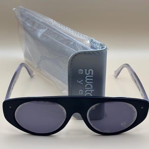 Puede incluir: Gafas de sol negras con forma de ojo de gato con lentes y patillas moradas, y un estuche a juego. El estuche es azul claro y tiene la inscripción "swatch eye".
