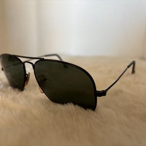 Pode incluir: &Oacute;culos de sol aviador pretos com lentes verde escuro. Os &oacute;culos t&ecirc;m uma ponte dupla e arma&ccedil;&otilde;es finas de metal preto. As lentes s&atilde;o ligeiramente refletivas. Os &oacute;culos de sol est&atilde;o sobre uma superf&iacute;cie branca e fofa.