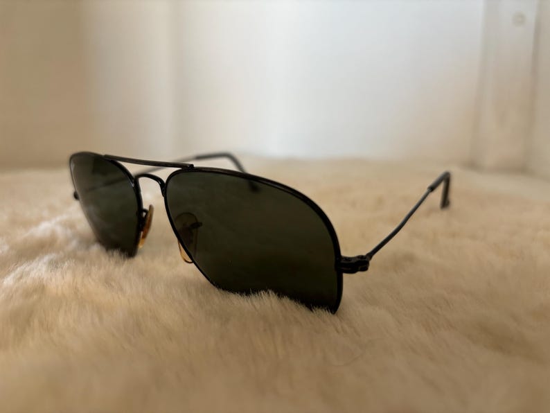Pode incluir: &Oacute;culos de sol aviador pretos com lentes verde escuro. Os &oacute;culos t&ecirc;m uma ponte dupla e arma&ccedil;&otilde;es finas de metal preto. As lentes s&atilde;o ligeiramente refletivas. Os &oacute;culos de sol est&atilde;o sobre uma superf&iacute;cie branca e fofa.