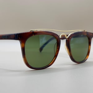 May include: Brown tortoiseshell sunglasses with green lenses and a gold-colored bridge. The frames have a classic, square shape. The sunglasses are a stylish accessory.