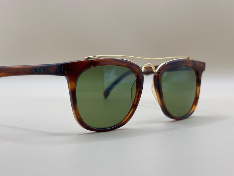 May include: Brown tortoiseshell sunglasses with green lenses and a gold-colored bridge. The frames have a classic, square shape. The sunglasses are a stylish accessory.