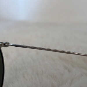 K&ouml;nnte beinhalten: Nahaufnahme einer silberfarbenen Sonnenbrille mit dunklen Gl&auml;sern. Der d&uuml;nne Metallrahmen tr&auml;gt den Aufdruck "Ray-Ban MADE IN ITALY" auf dem B&uuml;gel.