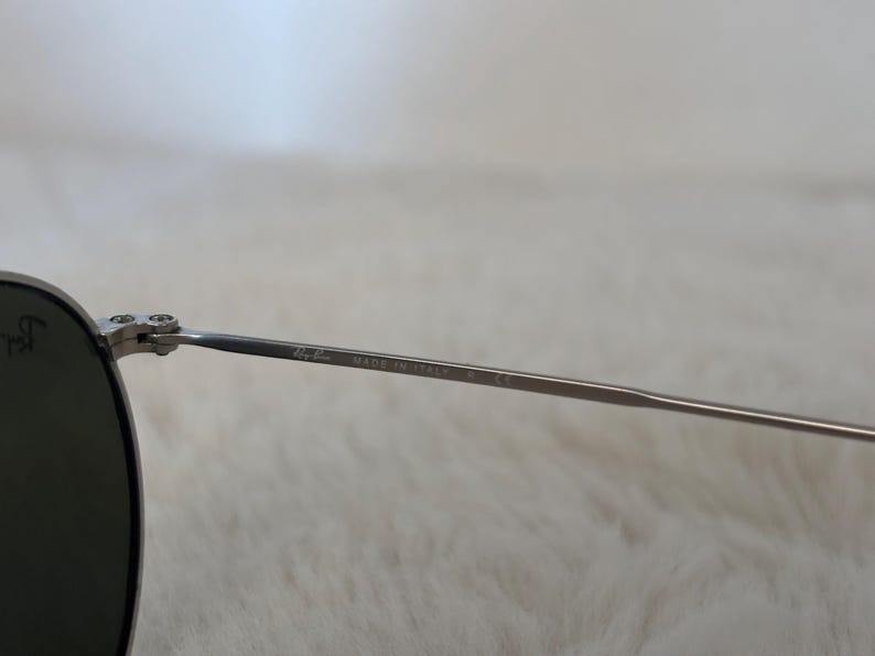 K&ouml;nnte beinhalten: Nahaufnahme einer silberfarbenen Sonnenbrille mit dunklen Gl&auml;sern. Der d&uuml;nne Metallrahmen tr&auml;gt den Aufdruck "Ray-Ban MADE IN ITALY" auf dem B&uuml;gel.