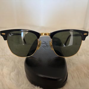 May include: Black and gold-rimmed sunglasses with green lenses. The sunglasses have a classic design with a black upper frame and gold accents. The Ray-Ban logo is visible on the lens and frame. The sunglasses are resting on a black case.