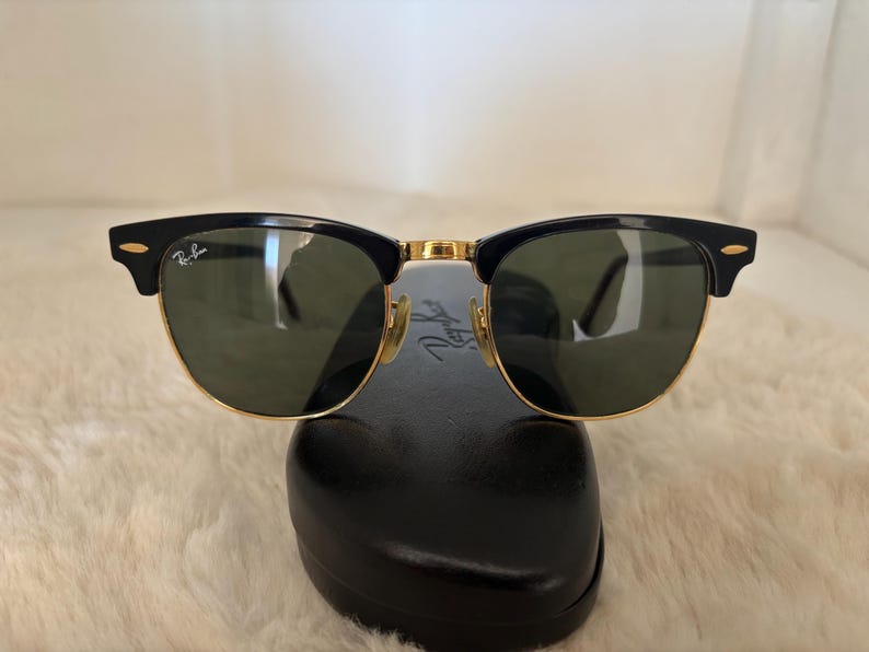 May include: Black and gold-rimmed sunglasses with green lenses. The sunglasses have a classic design with a black upper frame and gold accents. The Ray-Ban logo is visible on the lens and frame. The sunglasses are resting on a black case.