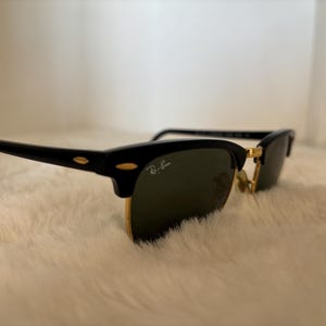 May include: Black and gold-rimmed sunglasses with dark lenses. The sunglasses feature a classic design with a black upper frame and temples, and gold-coloured lower rims. The Ray-Ban logo is visible on the lens.