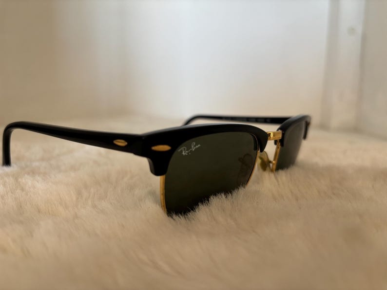 May include: Black and gold-rimmed sunglasses with dark lenses. The sunglasses feature a classic design with a black upper frame and temples, and gold-coloured lower rims. The Ray-Ban logo is visible on the lens.